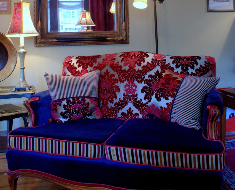 May include: A blue velvet sofa with a red and gold patterned upholstery. The sofa has two pillows, one with a striped pattern and the other with a floral pattern. The sofa is in a living room setting.