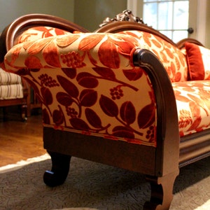 May include: A vintage-style chaise lounge with a dark wood frame and ornate detailing. The upholstery features a cream-colored background with a pattern of large, red-orange leaves and berries. A matching pillow rests on the seat.