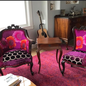 May include: Two antique armchairs with dark wood frames and vibrant purple floral patterned upholstery. The seats feature a patchwork design with a geometric pattern. A small wooden table and a guitar are in the background, set on a patterned rug.
