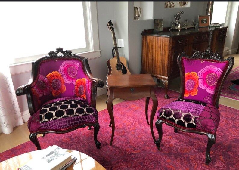 May include: Two antique armchairs with dark wood frames and vibrant purple floral patterned upholstery. The seats feature a patchwork design with a geometric pattern. A small wooden table and a guitar are in the background, set on a patterned rug.