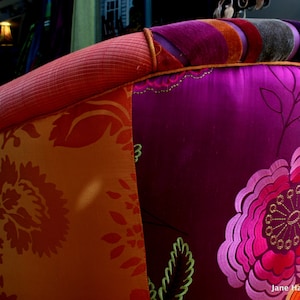 May include: Close-up of a colorful upholstered chair back. The chair features a vibrant mix of fabrics, including orange floral patterns, deep purple with embroidered flowers, and a striped top in various colors. The armrest is a textured coral color.