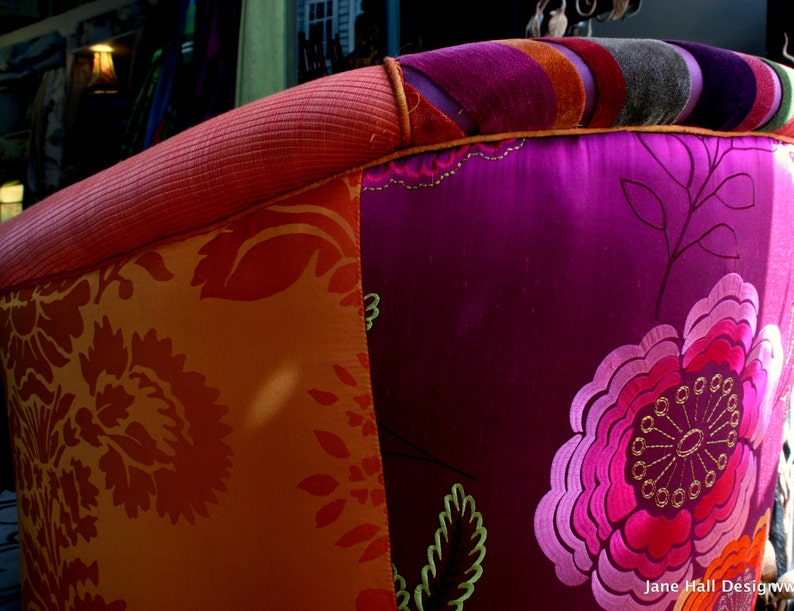 May include: Close-up of a colorful upholstered chair back. The chair features a vibrant mix of fabrics, including orange floral patterns, deep purple with embroidered flowers, and a striped top in various colors. The armrest is a textured coral color.
