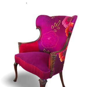 May include: Wingback chair with a high back and floral pattern in shades of pink, red, and purple. The chair has wooden arms and legs with carved details. The seat and back are upholstered in a plush fabric.
