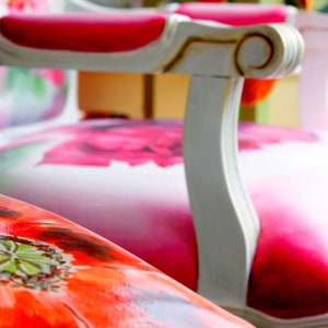 May include: Two white chairs with floral patterned upholstery. The chair in the foreground has a red poppy design, while the chair in the background has a pink floral design.