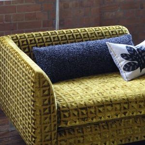 May include: A mustard-yellow sofa with a geometric pattern, accompanied by a dark blue bolster cushion and a decorative cushion with a black and white leaf design. The sofa is set against a brick wall.