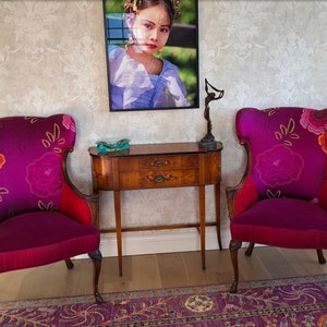 May include: Two ornate armchairs with high backs and floral patterns in shades of purple, pink, and orange. The chairs have wooden frames and are set against a patterned wallpaper. A small wooden table with drawers sits between them.
