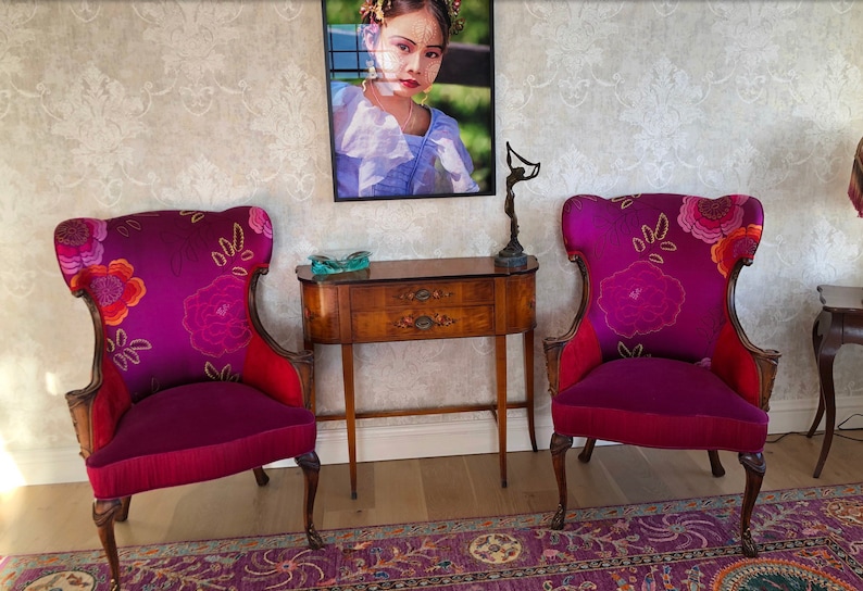 May include: Two ornate armchairs with high backs and floral patterns in shades of purple, pink, and orange. The chairs have wooden frames and are set against a patterned wallpaper. A small wooden table with drawers sits between them.