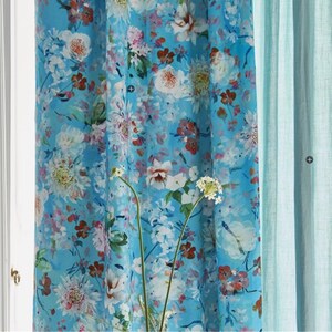 Pode incluir: Cortinas com estampa floral em tons de azul, branco, rosa e vermelho. As cortinas apresentam um padrão repetido de várias flores. Uma cortina azul claro lisa é visível à direita. As cortinas estão penduradas em frente a uma janela.
