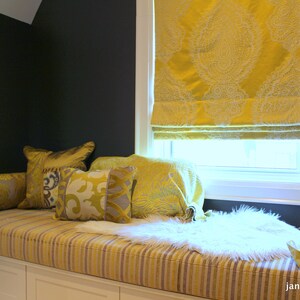 May include: A window seat with a white cushion and throw pillows in shades of yellow and gray. The window has a yellow patterned roman shade. The seat is upholstered in a striped fabric.
