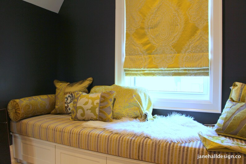 May include: A window seat with a white cushion and throw pillows in shades of yellow and gray. The window has a yellow patterned roman shade. The seat is upholstered in a striped fabric.