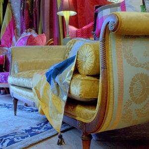 May include: A vintage-style yellow velvet chaise lounge with a curved wooden frame and decorative floral patterned fabric. The chaise features a bolster pillow and is draped with a blue and yellow fabric with a tassel.