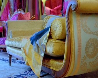 May include: A vintage-style yellow velvet chaise lounge with a curved wooden frame and decorative floral patterned fabric. The chaise features a bolster pillow and is draped with a blue and yellow fabric with a tassel.