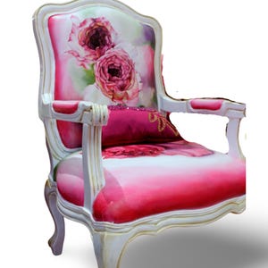 May include: A white and gold armchair with a pink and white floral patterned upholstery. The chair has a curved back and a padded seat with a pink pillow.