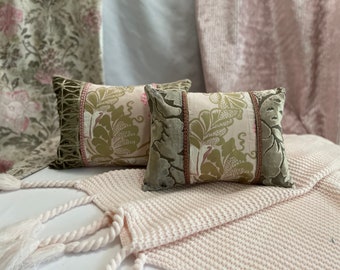 May include: Two decorative throw pillows with a floral pattern in shades of green, pink, and white. The pillows have a velvet trim and are sitting on a white knitted blanket.