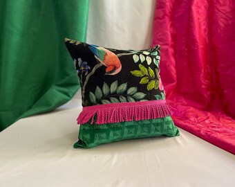 May include: A black pillow with a colorful bird and floral print. The pillow has a pink fringe trim and a green velvet back.