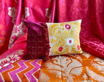 May include: A white pillow with gold embroidery of a floral design, resting on a pink and orange patterned fabric. The pillow has a pink and red trim with a tassel.