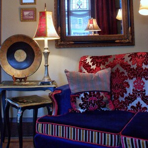 May include: A blue velvet sofa with red and gold patterned cushions. The sofa has a gold and black striped trim. A lamp with a red and white lampshade sits on a black table next to the sofa.