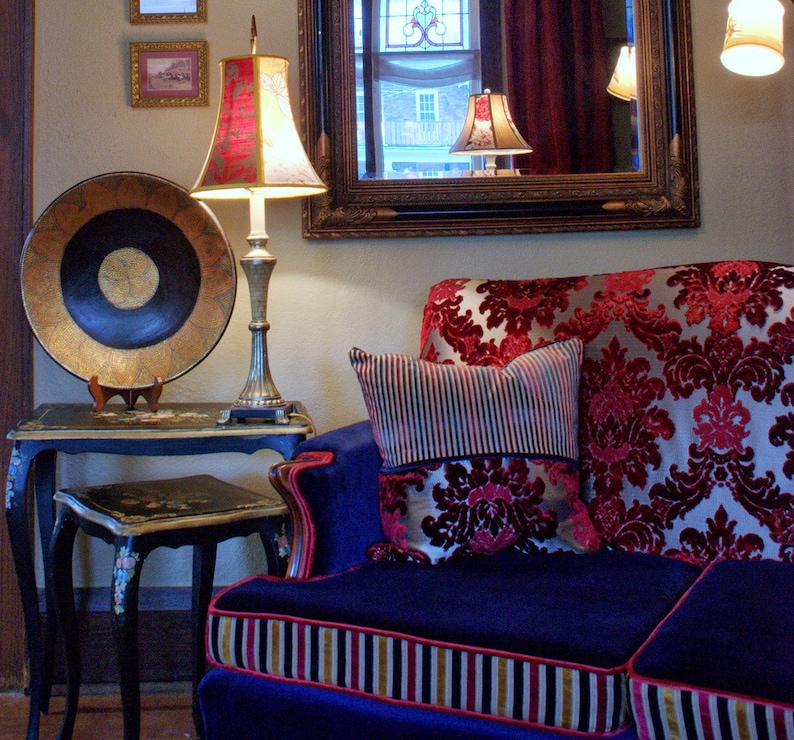 May include: A blue velvet sofa with red and gold patterned cushions. The sofa has a gold and black striped trim. A lamp with a red and white lampshade sits on a black table next to the sofa.