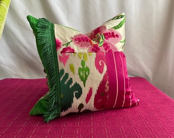 May include: A decorative pillow with a patchwork design featuring pink and green floral embroidery, a white and green ikat fabric, and a pink and white striped fabric. The pillow has a green fringe trim.