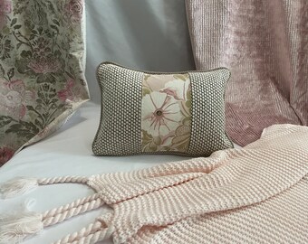 May include: A soft, pink knitted throw blanket with tassels on the ends. The blanket is draped over a white surface with a decorative pillow featuring a floral pattern in shades of pink and green.