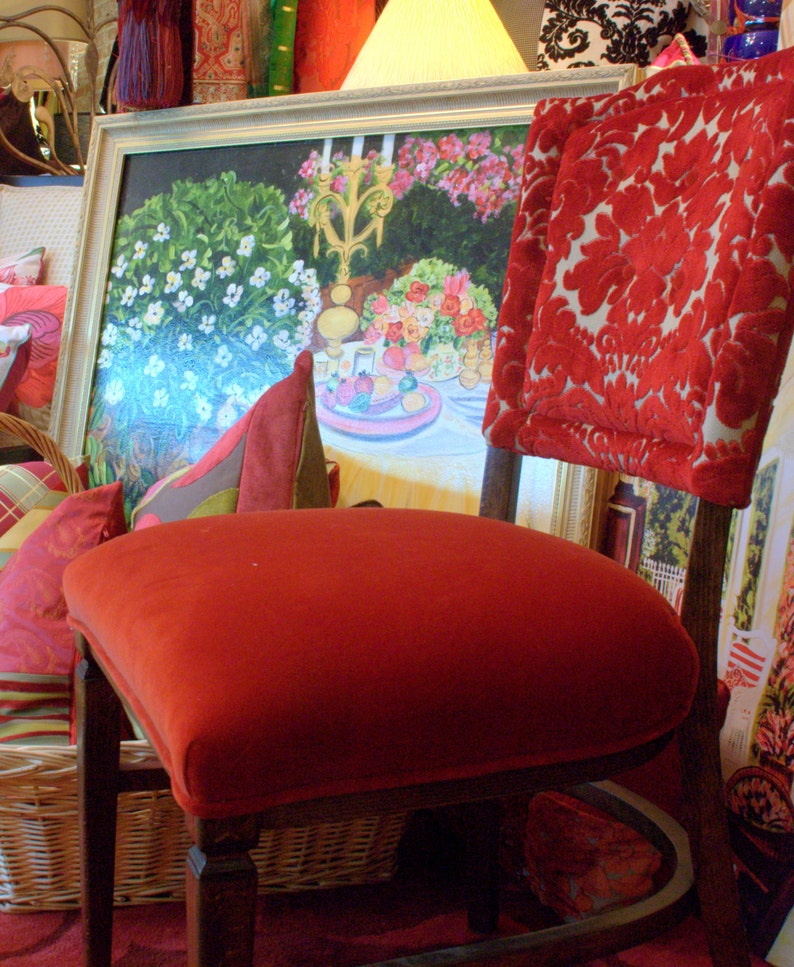 May include: A red velvet chair with a floral painting in a gold frame behind it. The chair has a red velvet seat and back, and the painting depicts a still life with flowers and fruit.