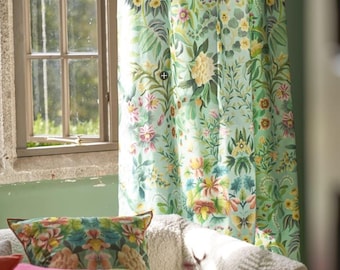 May include: A pale blue floral curtain with a matching cushion on a white sofa. The curtain displays a pattern of colourful flowers and green leaves. A red cushion is also on the sofa. A window is visible in the background.