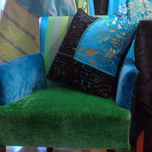 Op de afbeelding: Een groene fluwelen fauteuil met een blauwe fluwelen armleuning en een blauw-zwart gepatroneerd kussen. Het kussen heeft een geometrisch ontwerp met gouden accenten.