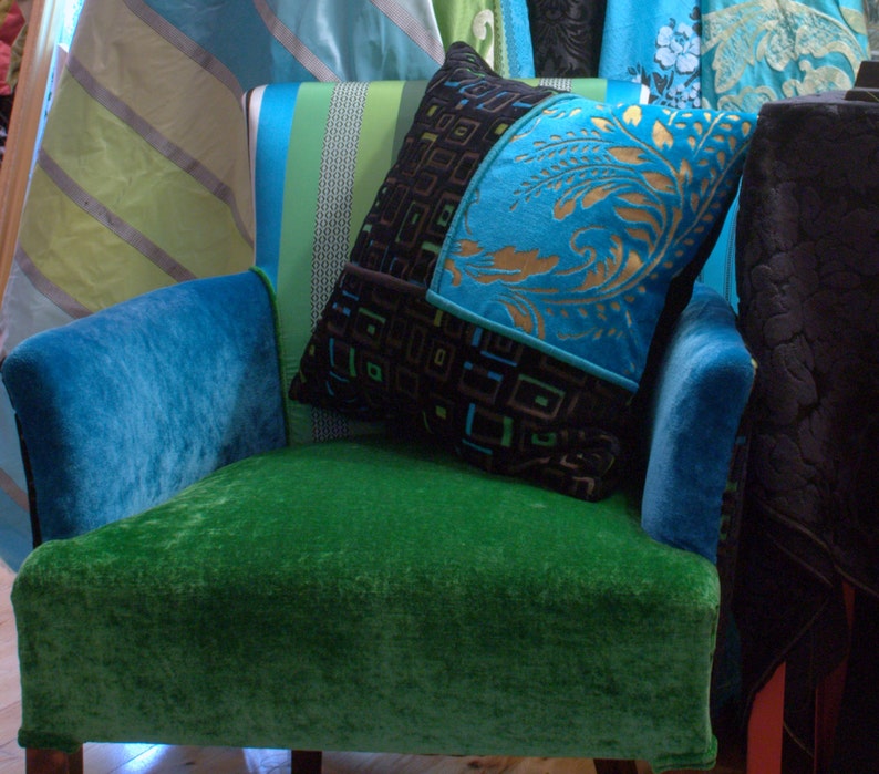 Op de afbeelding: Een groene fluwelen fauteuil met een blauwe fluwelen armleuning en een blauw-zwart gepatroneerd kussen. Het kussen heeft een geometrisch ontwerp met gouden accenten.