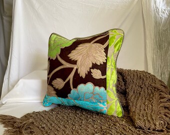 Può includere: Cuscino quadrato decorativo con motivo floreale. Presenta uno sfondo marrone scuro con foglie beige e verdi e un accento floreale turchese. I bordi sono rifiniti con velluto verde e un cordoncino nero.