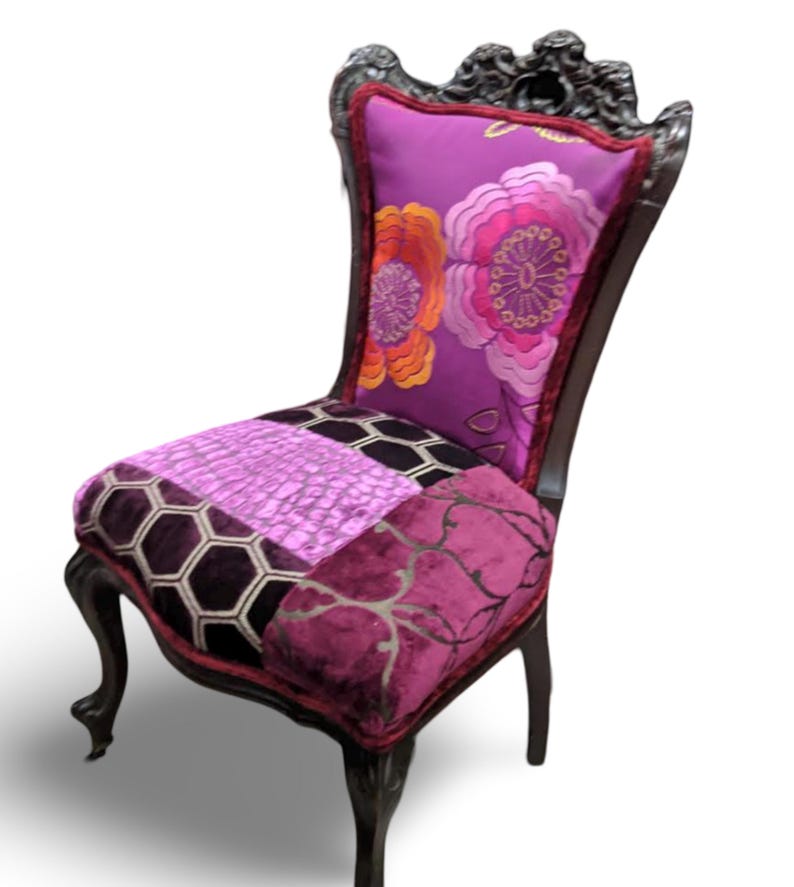 May include: A decorative chair with a dark wood frame and ornate carvings. The backrest features a purple floral pattern, while the seat is a patchwork of various purple fabrics. The chair has curved legs and a red trim.