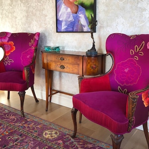 May include: Two ornate armchairs with a vibrant floral pattern on a deep purple background. The chairs have wooden frames, red accents, and are placed in a room with a patterned rug and wallpaper. A small wooden table is between the chairs.