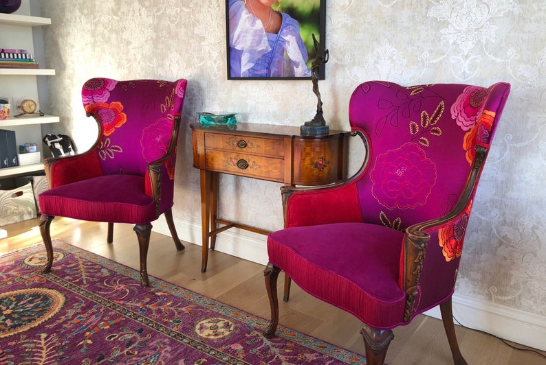 May include: Two ornate armchairs with a vibrant floral pattern on a deep purple background. The chairs have wooden frames, red accents, and are placed in a room with a patterned rug and wallpaper. A small wooden table is between the chairs.