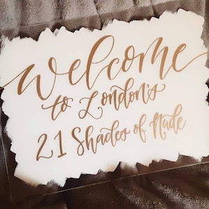 May include: A clear acrylic sign with gold calligraphy text that reads "Welcome to London's 21 Shades of Nude." The text is on a white, brushstroke-style background.