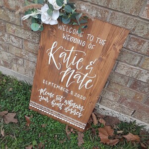 May include: A wooden wedding sign with white calligraphy that reads "Welcome to the wedding of Katie & Nate September 5, 2020 Please sit wherever your heart desires". The sign is decorated with a white floral arrangement.