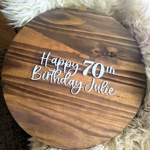 May include: A round wooden sign with the text "Happy 70th Birthday Julie" in white lettering.