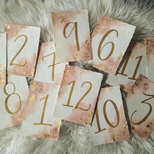 May include: A collection of handmade table numbers. Each card features a unique number in gold script, set against a watercolor wash of blush pink and gold leaf accents. Ideal for weddings or events.