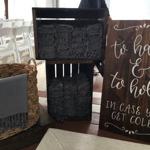 May include: A wooden sign with white lettering that reads "to have & to hold in case you get cold".  The sign is propped up against a stack of gray blankets in wooden crates.