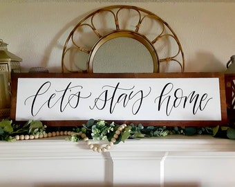 May include: A white wooden sign with a brown frame, featuring the text "Let's Stay Home" in black calligraphy. The sign is displayed on a white mantelpiece with greenery and wooden beads.