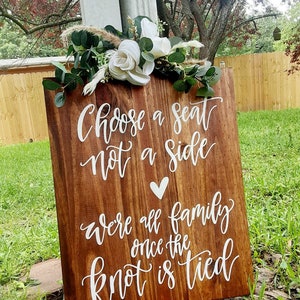 May include: A wooden sign with white calligraphy that reads "Choose a seat, not a side. We're all family once the knot is tied." The sign is decorated with white flowers and greenery.