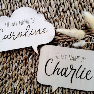 May include: Two wooden speech bubble signs with the text "Hi, my name is Caroline" and "Hi, my name is Charlie" written in black.