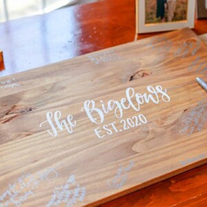 May include: A wooden guest book with the text "The Bigelows EST. 2020" in white lettering. The book is on a wooden table and has several signatures in silver pen.