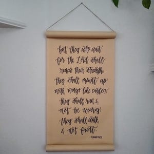 May include: A tan scroll with black calligraphy script, hanging from a thin wire. The text reads, "but they who wait for the Lord shall renew their strength..." and is from Isaiah 40:31. The scroll is mounted on wooden dowels.