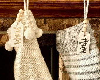 May include: Two knitted Christmas stockings with personalized tags. One stocking is cream-colored with white pom-poms and a tag that says "Jennifer". The other is gray and white striped with a tag that says "Mason". Both are hanging from a wooden mantel.