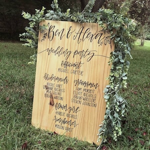 May include: A wooden sign with the names "Ben & Alexa" and the text "wedding party" written in black calligraphy. The sign lists the officiant, bridesmaids, groomsmen, flower girl, and ring bearer.