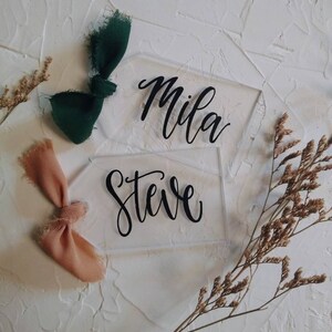 May include: Two clear acrylic place cards with the names "Mila" and "Steve" written in black calligraphy. The cards are decorated with a green and a peach ribbon.