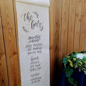 May include: A white paper scroll with black calligraphy listing the drinks available at a bar. The drinks include specialty cocktails, beer, and wine. The text reads: "the bar", "Specialty cocktails", "Dirty martini", "Old fashioned", "Moscow mule", "Beer", "Shiner", "Doo equio", "Miller lite", "Wine", "Chardonnay", "Pinot noir".