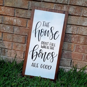 May include: A rectangular wooden sign with a white background and dark brown frame. The sign features black cursive text that reads, "THE house DON'T FALL WHEN THE bones ARE GOOD."