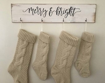 May include: A wooden sign with the words "merry & bright" in cursive script hangs above four cream-colored knitted Christmas stockings. The stockings are suspended by hooks, creating a festive holiday display.
