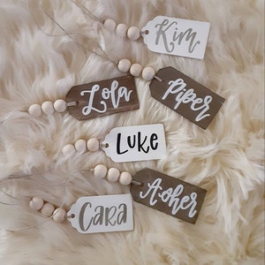 May include: Wooden name tags with names like "Kim", "Lola", "Piper", "Luke", "Cara", and "Asher" are displayed. The tags are in various colors, including white and brown, and are strung together with wooden beads. The names are written in a cursive font.