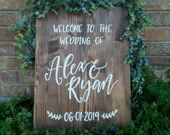 May include: A rustic wooden sign with greenery on the sides. The sign says "Welcome to the wedding of Alex & Ryan 06.01.2019".